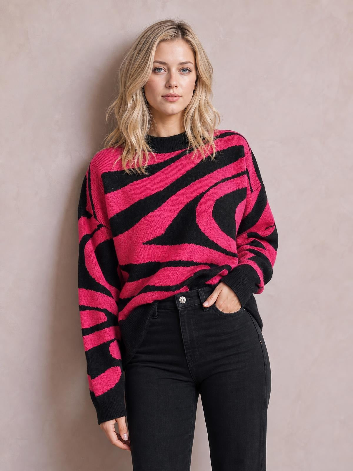 Model wears an oversized pink and black zebra print round neck sweater with long sleeves, ribbed cuffs, and a relaxed silhouette, posing with one hand in her jeans pocket.