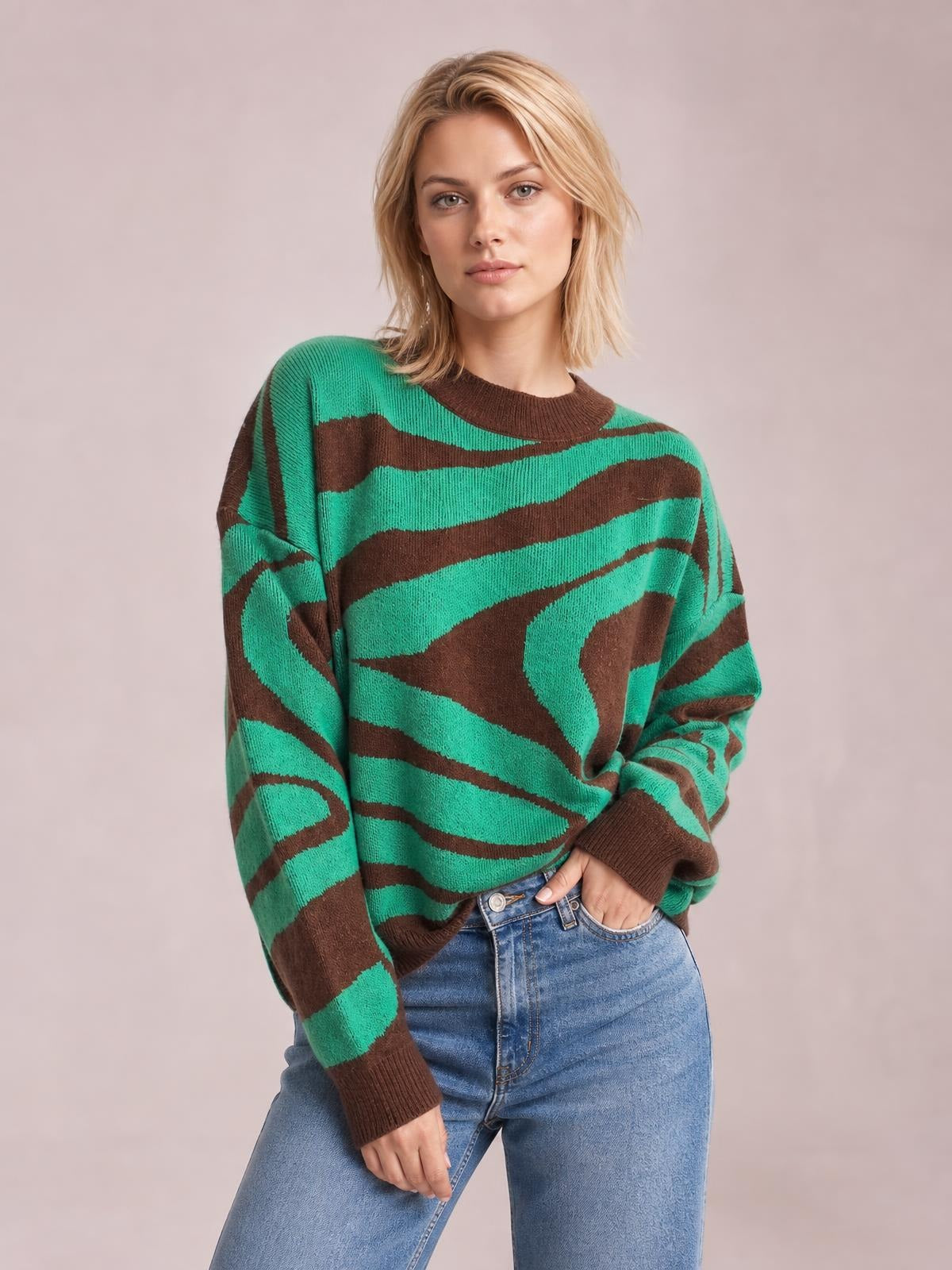 Model wearing an oversized green and brown zebra print round neck sweater with dropped shoulders, long sleeves, and ribbed cuffs, posing with one hand in her jeans pocket.