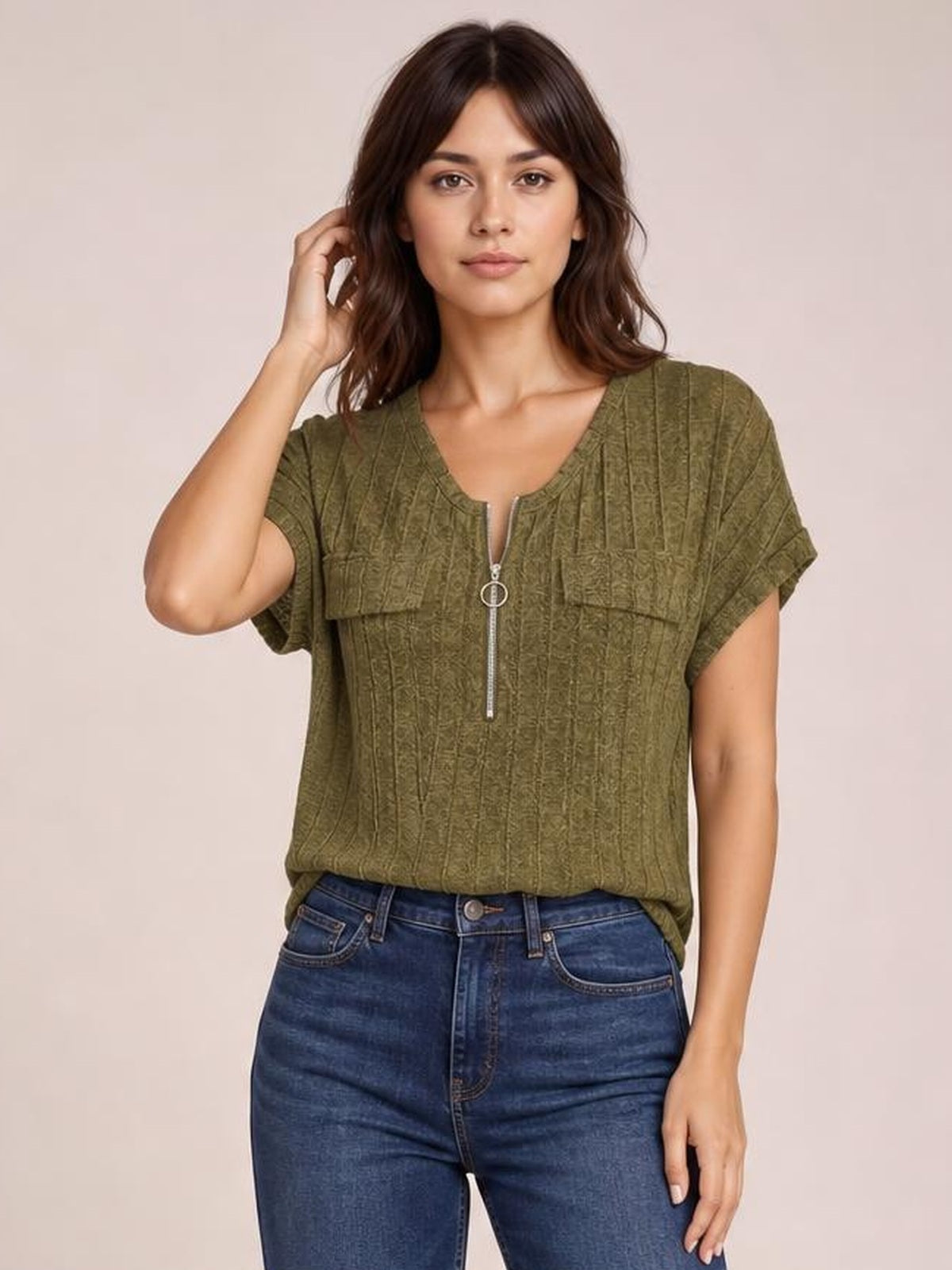 Model wearing an olive green ribbed short-sleeve T-shirt with a relaxed silhouette, zip-front closure, and rolled sleeve cuffs, posing with one hand in her hair.