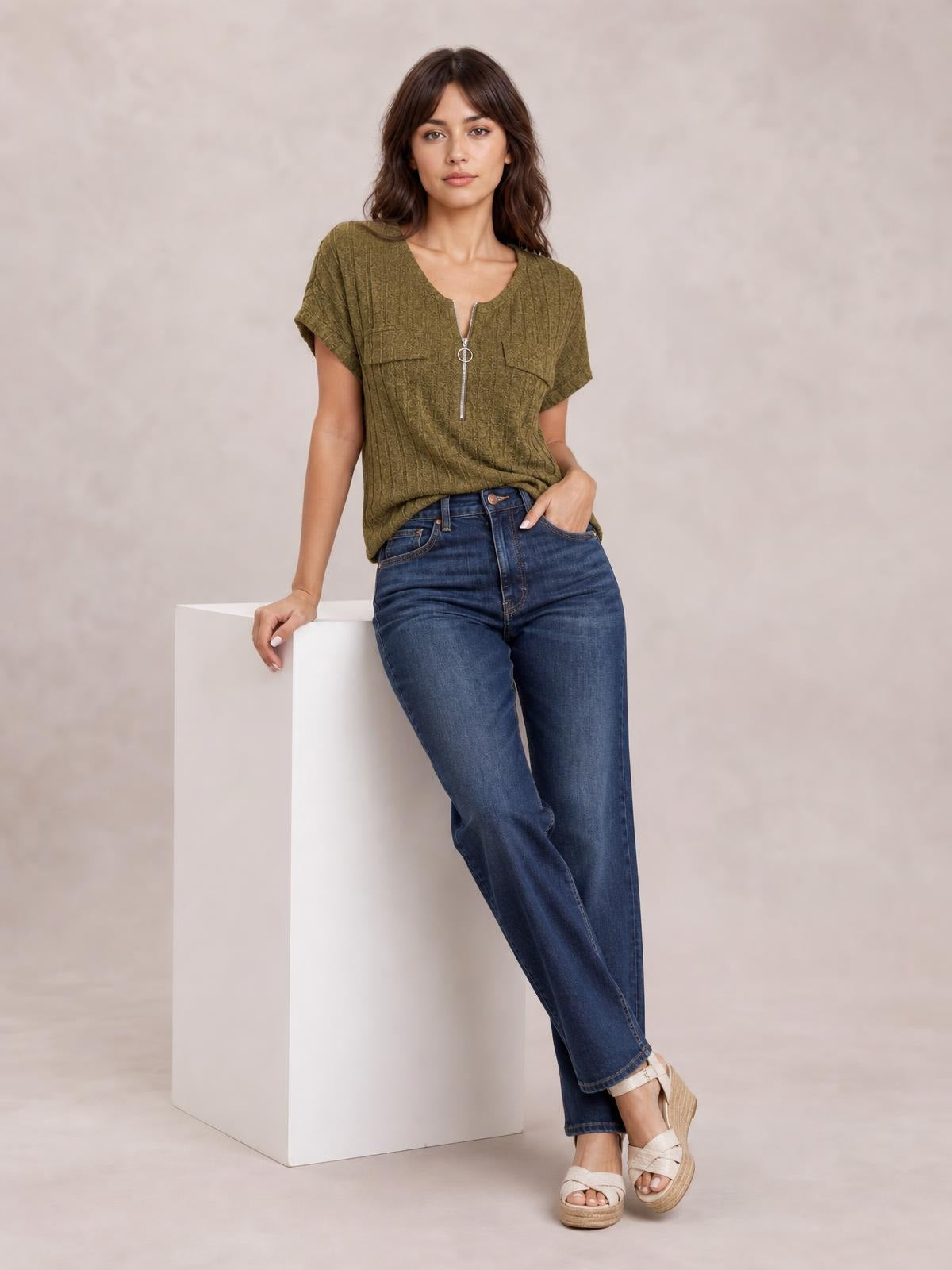 A model leans against a pedestal wearing an olive ribbed short-sleeve T-shirt with a relaxed silhouette, zip-front neckline, and chest pockets.