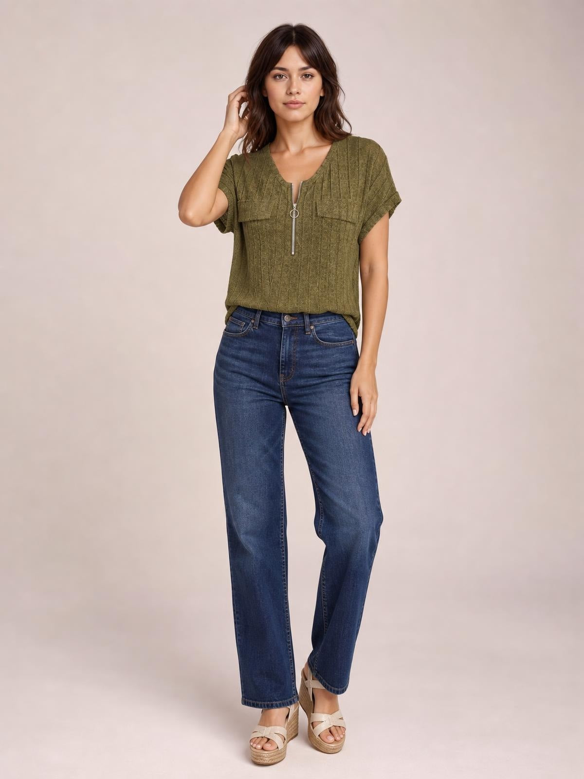 Model wearing an olive ribbed short sleeve T-shirt with a zip-front neckline and relaxed silhouette, standing with one hand touching her hair.