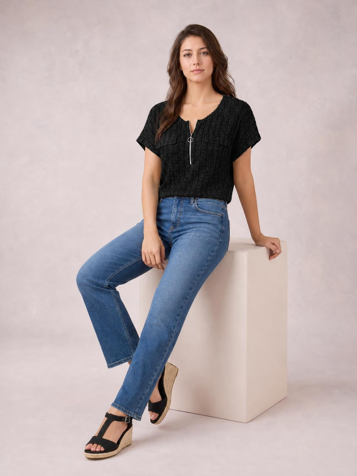 A seated model wears a black ribbed zip-front short sleeve T-shirt with a slightly relaxed silhouette and ring-pull zipper, paired with blue jeans.
