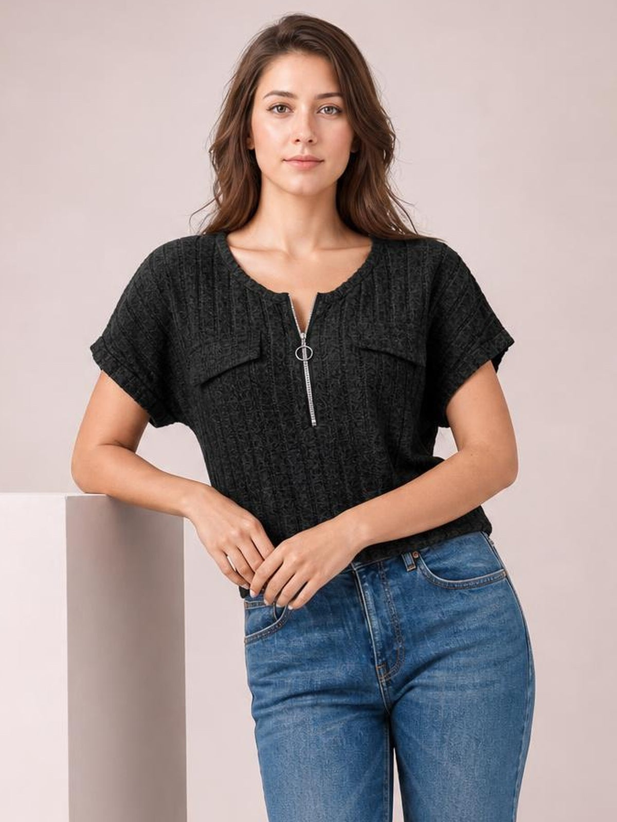A model wears a black ribbed short-sleeve T-shirt with a zip front and ring pull, relaxed silhouette, and visible flap chest pockets while standing with hands at her waist.