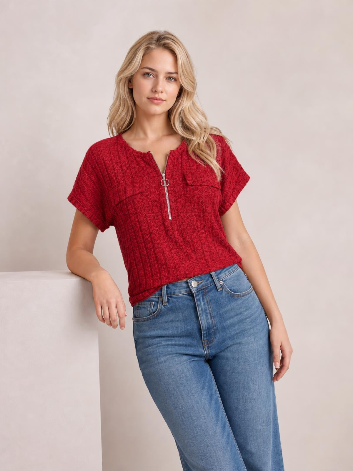 Model leans against a surface wearing a red ribbed short-sleeve T-shirt with a front zipper, relaxed silhouette, and wide sleeve cuffs.