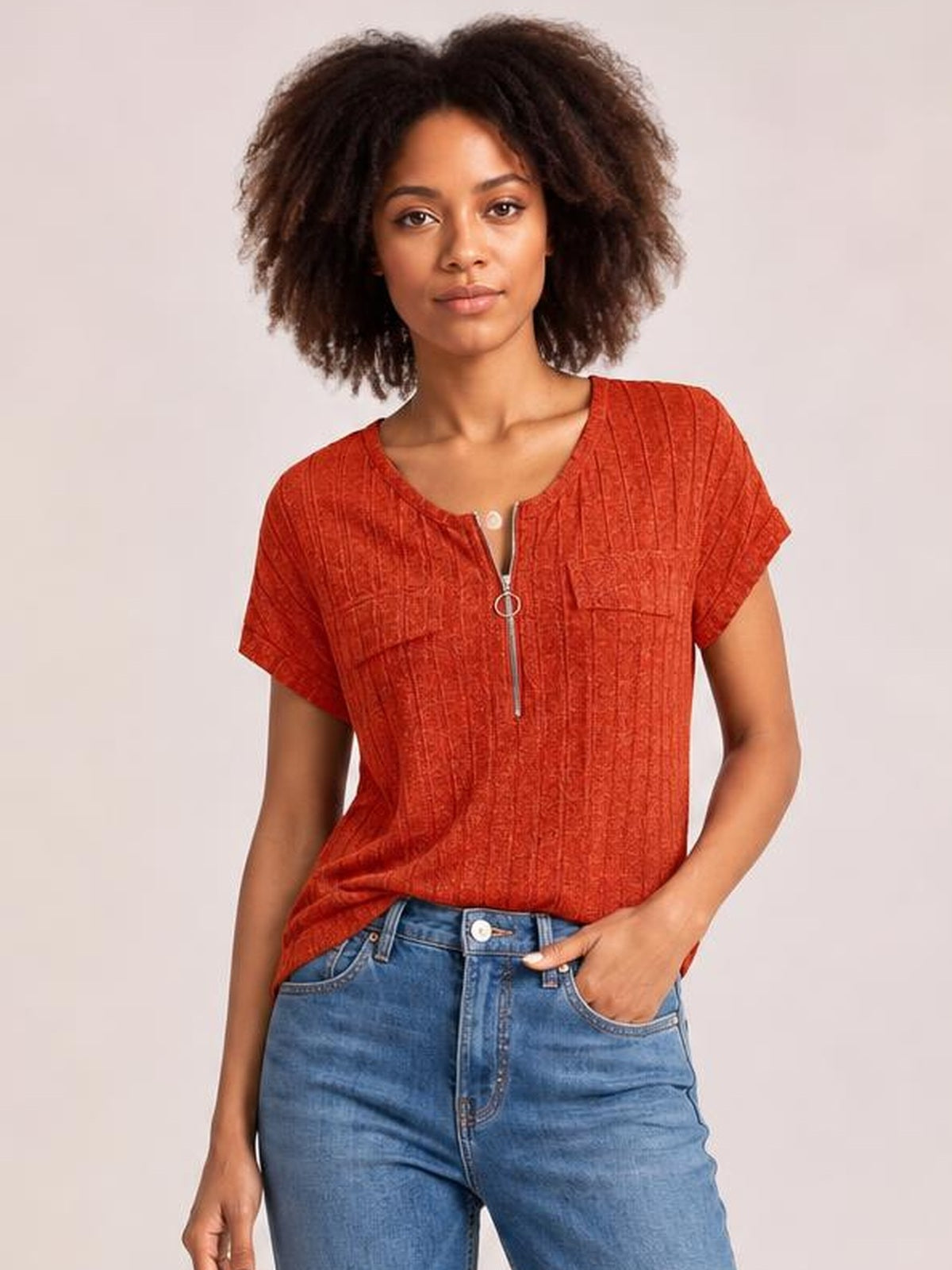 Model wearing a rust-red ribbed short sleeve T-shirt with a relaxed silhouette, round neckline, and front zipper, posing with one hand in her jeans pocket.