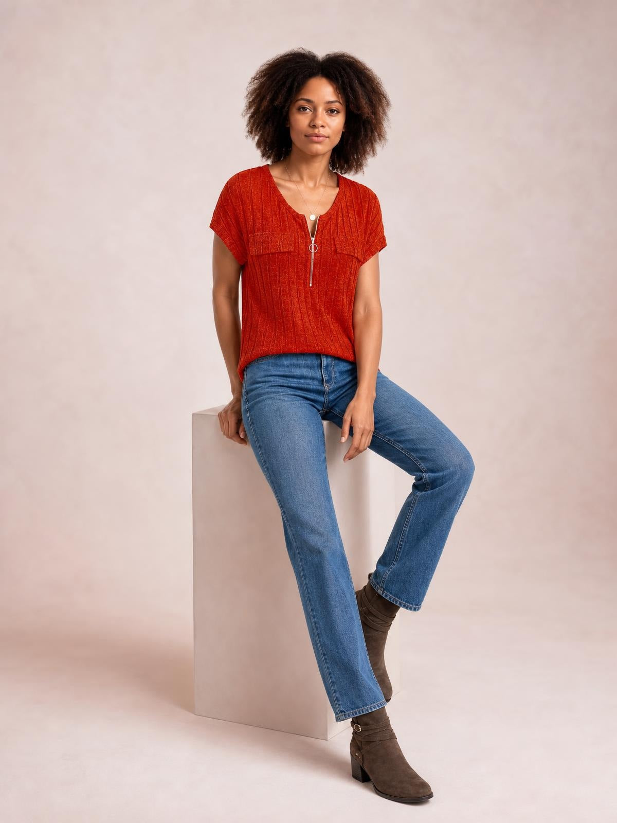 A seated model wears a rust-red ribbed short-sleeve T-shirt with a zip front, V-neckline, and relaxed straight silhouette.