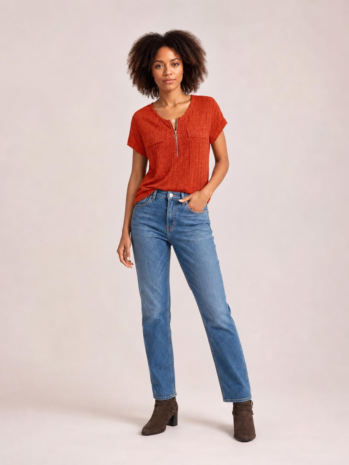 Model wears a rust-red ribbed short-sleeve T-shirt with a scoop neck, zip-front detail, and relaxed silhouette, posing with one hand in her jeans pocket.