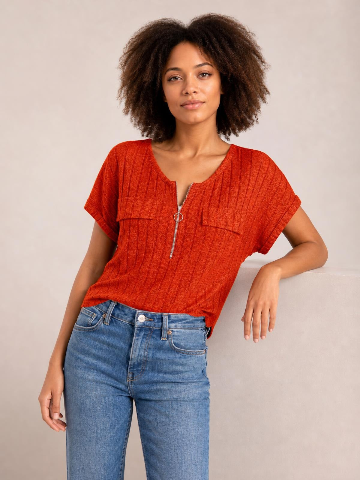 A model wears a rust-orange ribbed zip-front short sleeve T-shirt with a relaxed silhouette, V-neck opening, and visible chest pockets while posing with one arm resting.
