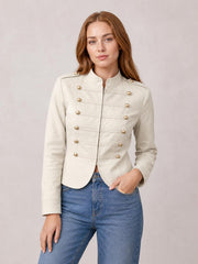 A model wears a cream cropped jacket with a zip front, stand collar, gold-tone buttons, long sleeves, and a fitted silhouette, posing with one hand in her pocket.