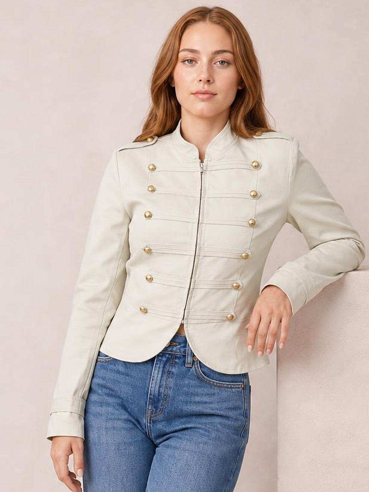 Model wears a cream cropped jacket with a front zip, high collar, long sleeves, gold button detailing, and a fitted silhouette while standing with one hand resting.