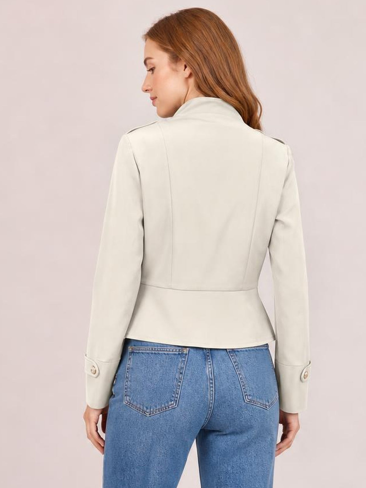 Model turned away in a cream cropped jacket with a structured back, high collar, long sleeves, buttoned cuffs, and a fitted waist-length silhouette.