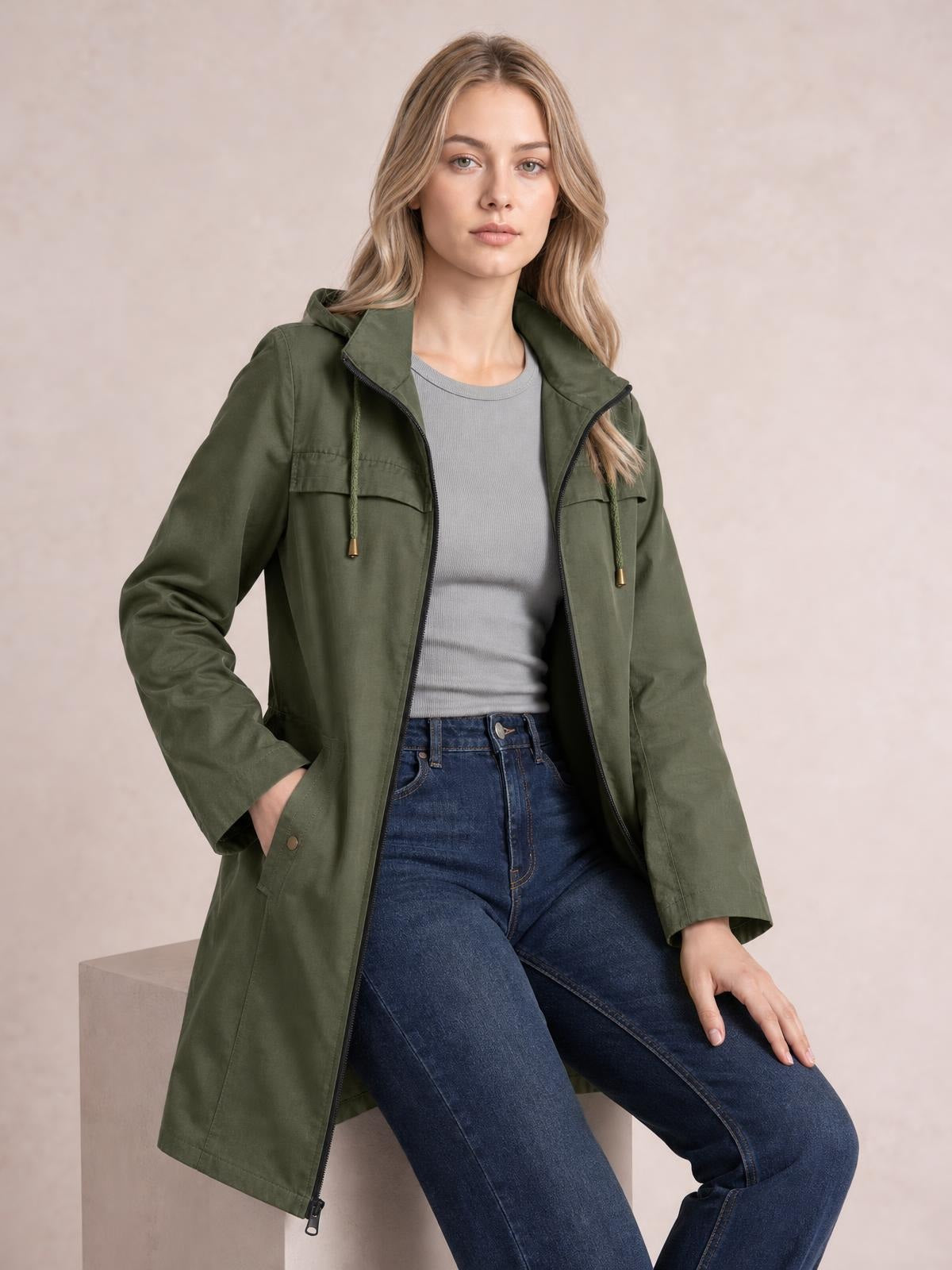 A woman poses seated in an olive green hooded jacket with an open zip front, drawstring hood, flap pockets, long regular silhouette, and relaxed sleeves.
