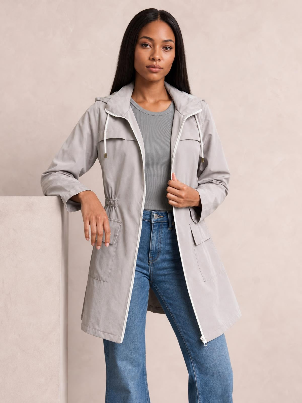 A model wears a light gray zip-up hooded jacket with a drawstring waist, long regular silhouette, flap pockets, and relaxed sleeves over a gray top and jeans.