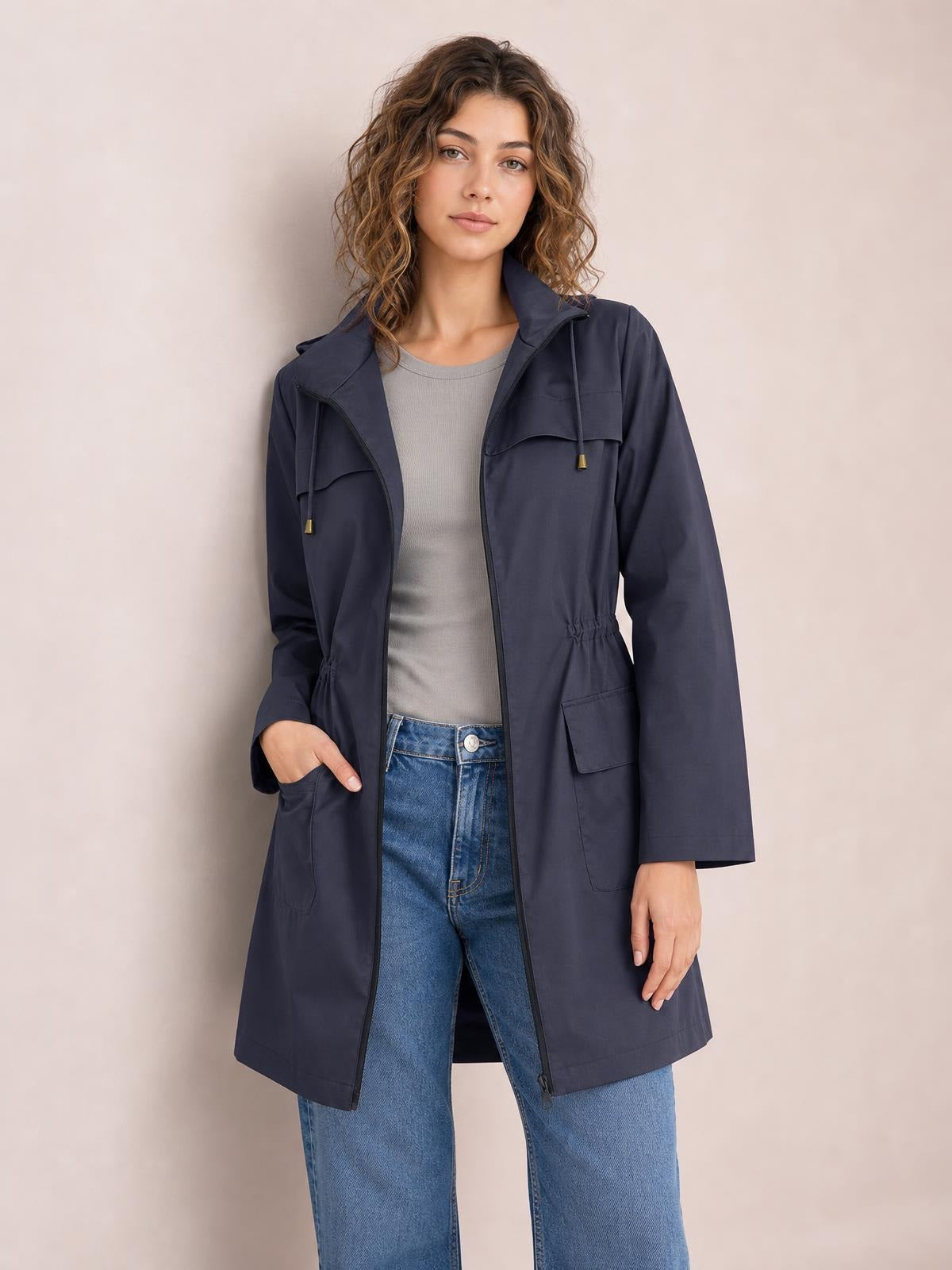 A standing model wears a navy hooded jacket with a front zip, drawstring waist, long regular-fit silhouette, flap chest and lower pockets, and long sleeves.