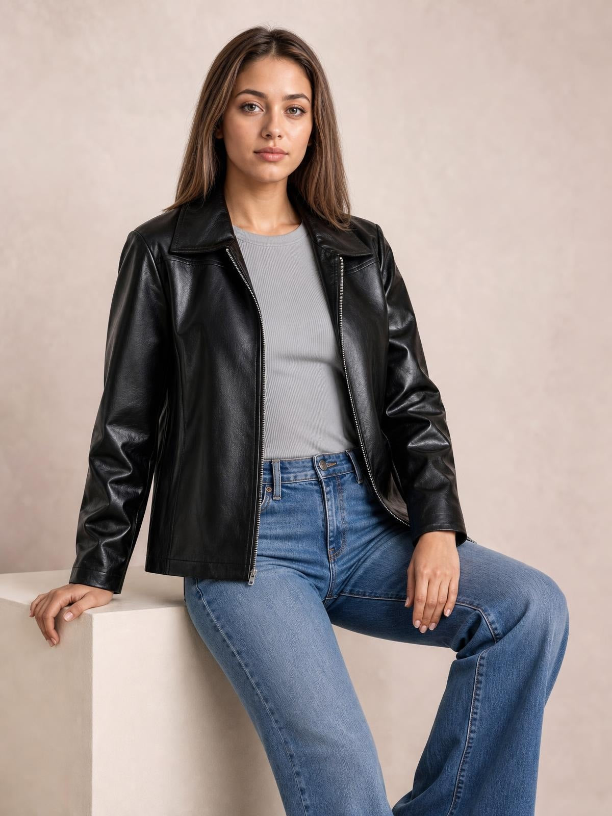 A woman sits wearing a black leather-look zip-up jacket with a wide collar, long sleeves, open front, and relaxed straight silhouette.