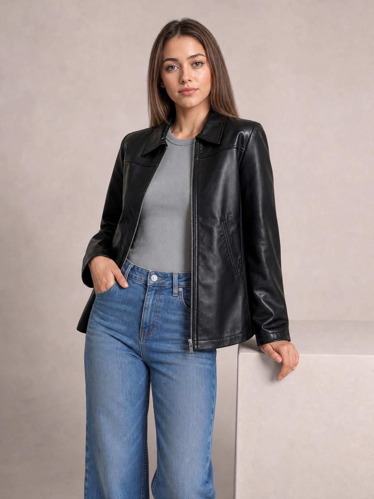 Model wearing a black zip-up leather jacket with a collared, pocketed design and relaxed silhouette, standing with one hand in her jeans pocket.