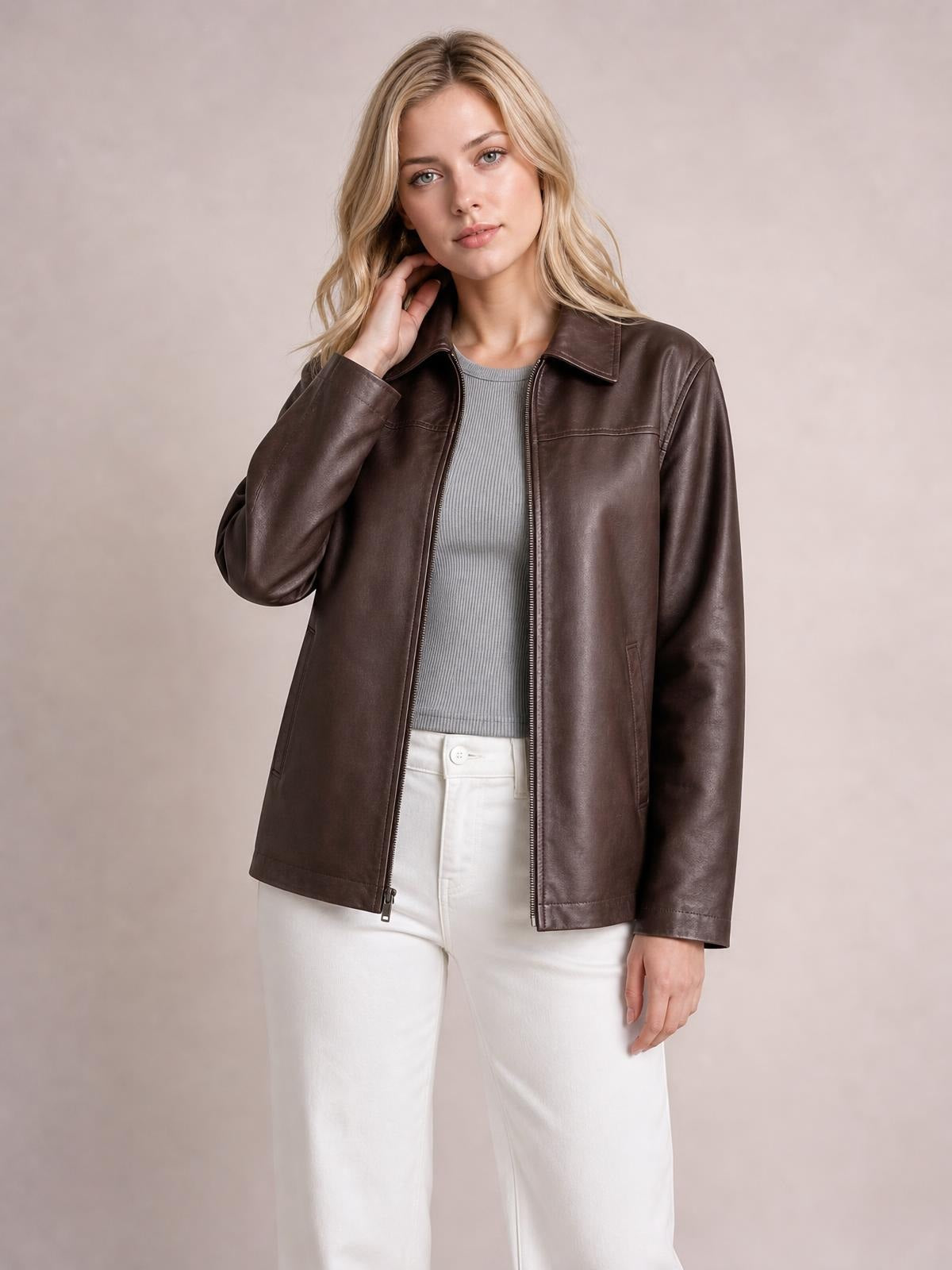 A model poses with one hand at her neck, wearing a brown leather-look zip-up jacket with a collared neckline, long sleeves, pockets, and a straight silhouette.