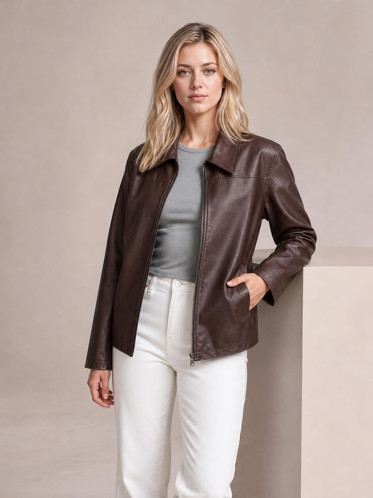 A model wears a brown leather jacket with a zip front, notched collar, long sleeves, pocketed construction, and a relaxed straight silhouette, posing with one hand in a pocket.