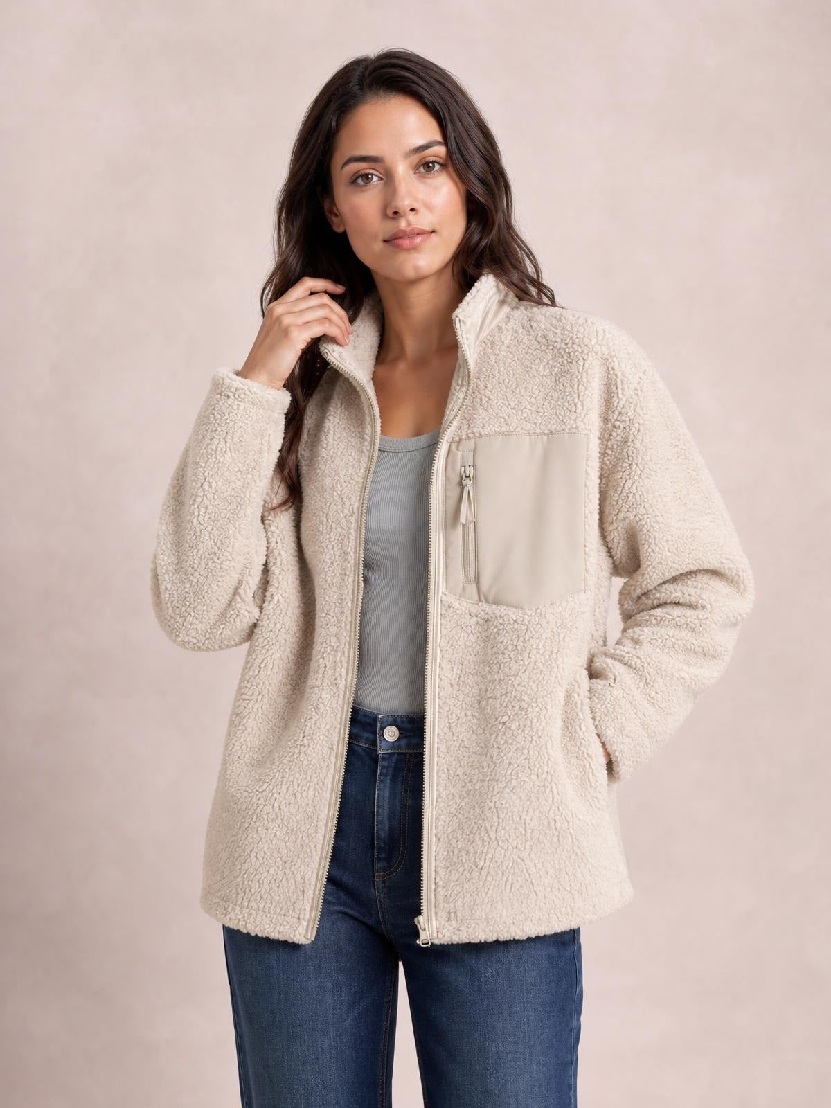 A model poses in a cream plush jacket with long sleeves, a full front zip, stand collar, relaxed silhouette, and a large zippered chest pocket.