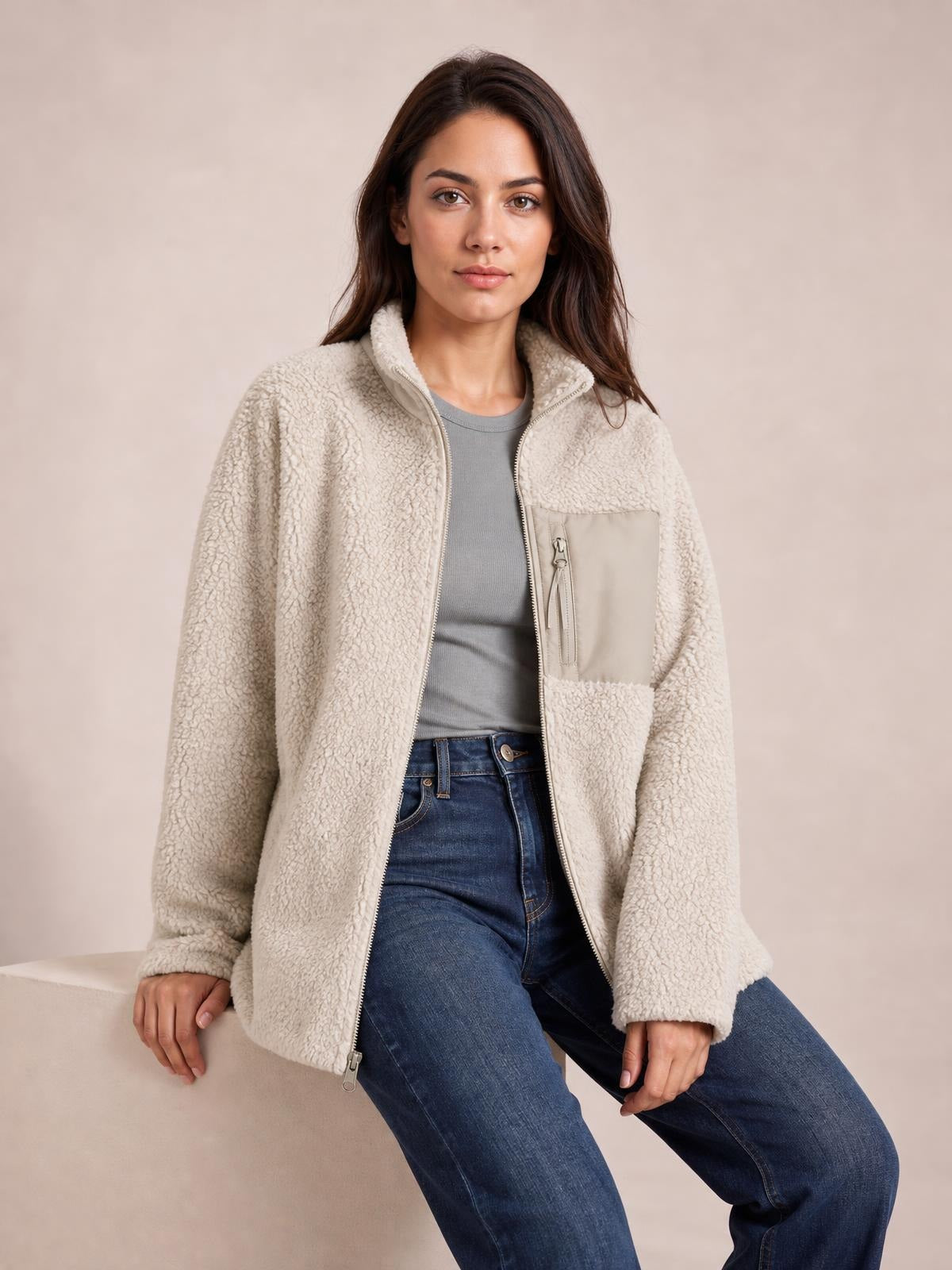 A seated model wears a cream plush jacket with long sleeves, a front zip closure, stand collar, zip chest pocket, and relaxed silhouette.