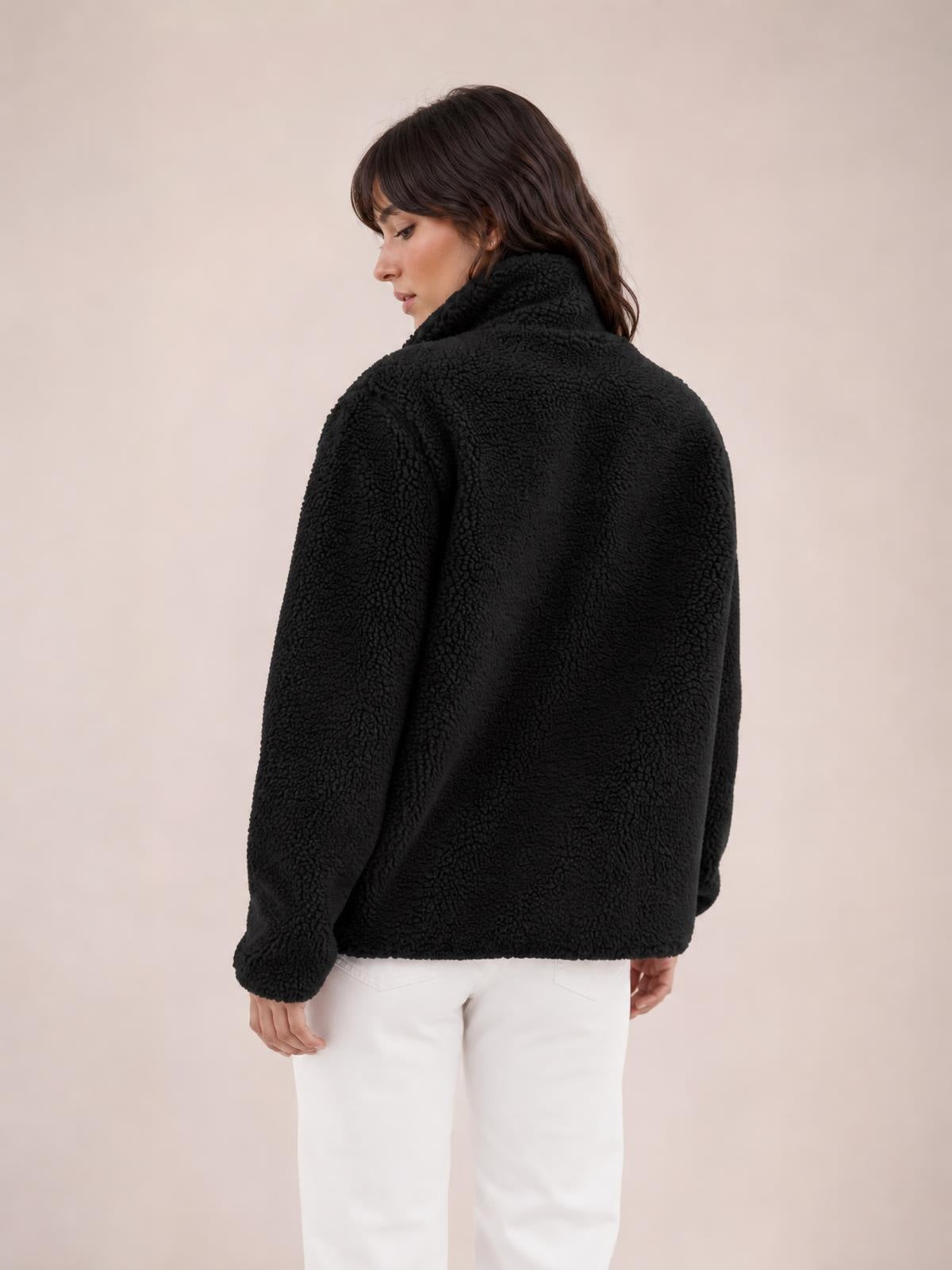 Model shown from the back in a black plush jacket with long sleeves and a high collar, standing in a relaxed, slightly turned pose with a boxy silhouette.