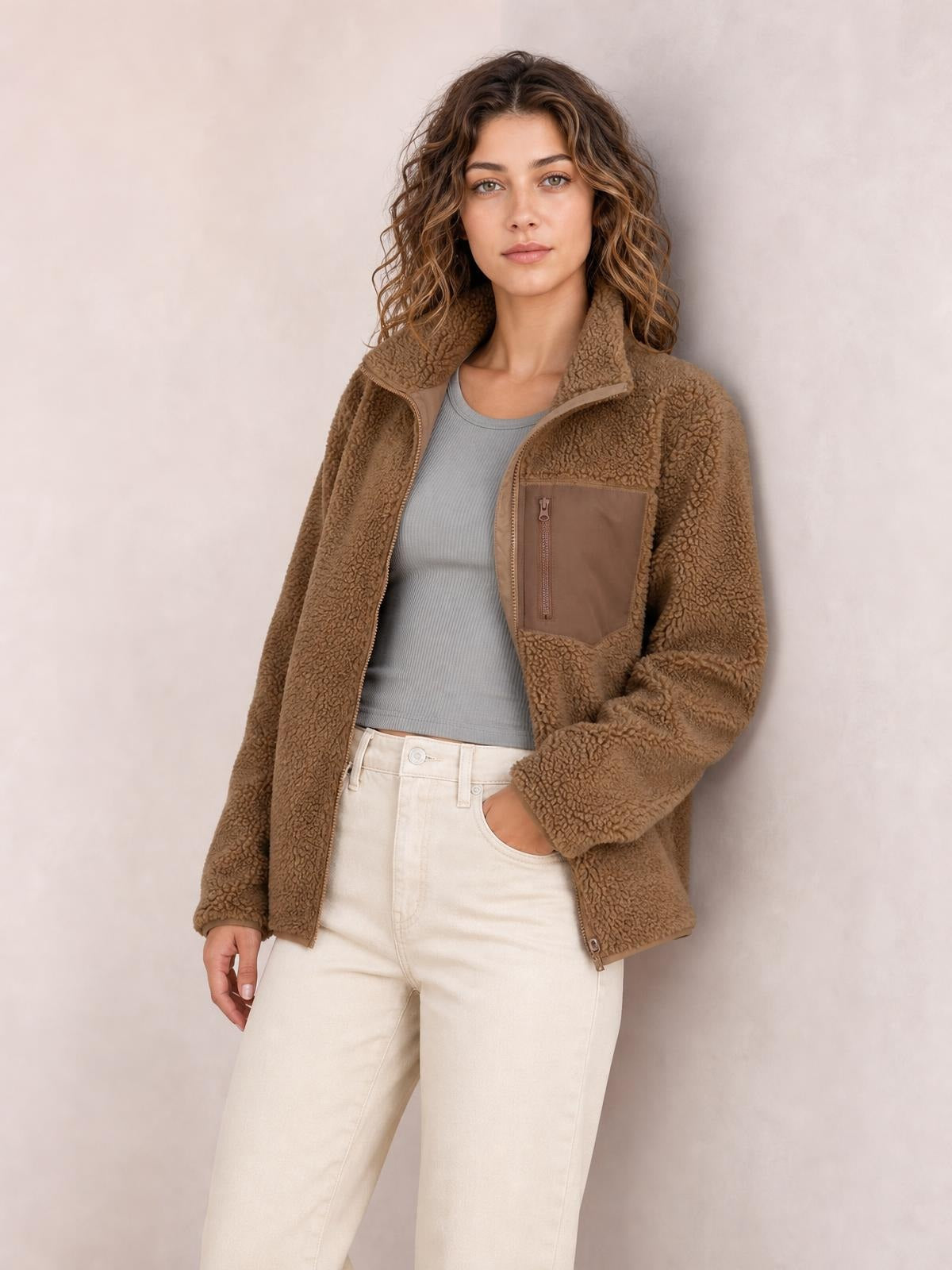 Model poses in a brown plush jacket with long sleeves, zip front, high collar, relaxed silhouette, and a visible zippered chest pocket.