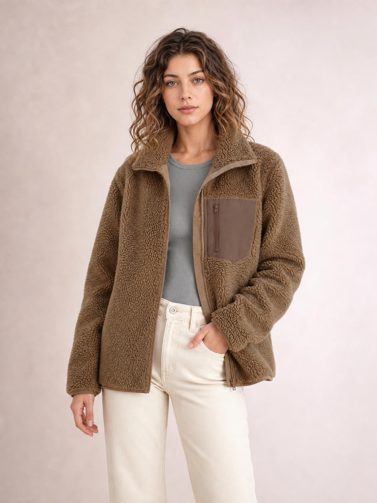 A model poses in a taupe plush jacket with long sleeves, a zip front, stand collar, relaxed silhouette, and visible zippered chest and side pockets.