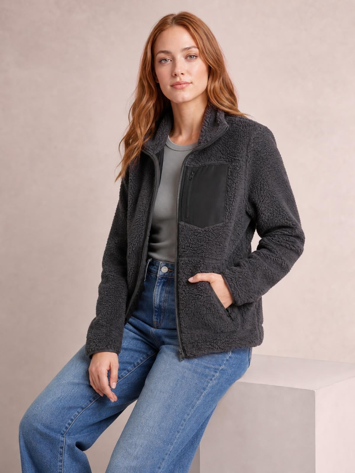 A seated model wears a charcoal plush jacket with long sleeves, front zip closure, stand collar, zippered chest pocket, and side pockets in a relaxed silhouette.