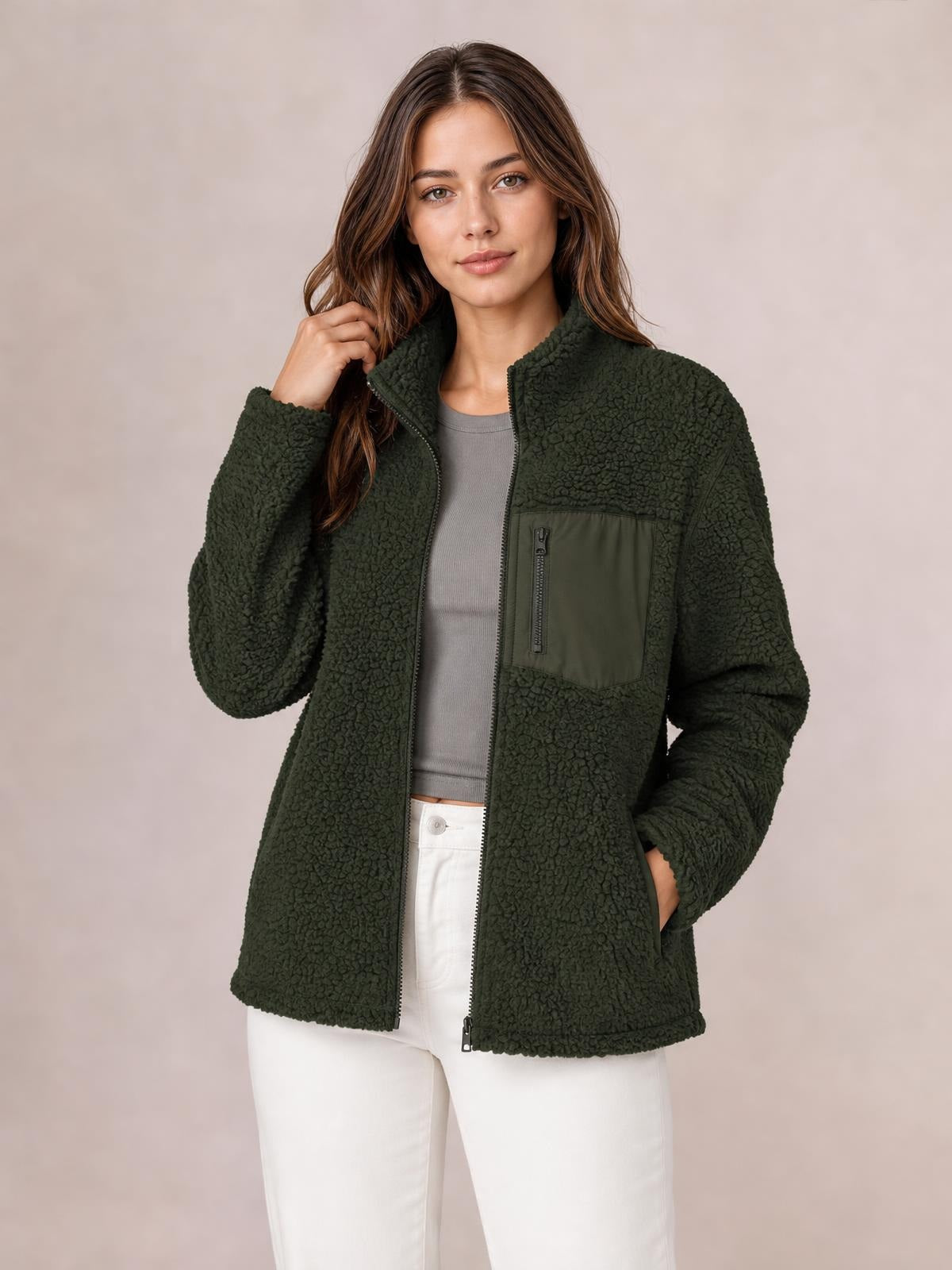 Model poses in a dark olive plush jacket with a zip front, long sleeves, stand collar, contrasting chest zip pocket, and side pockets.