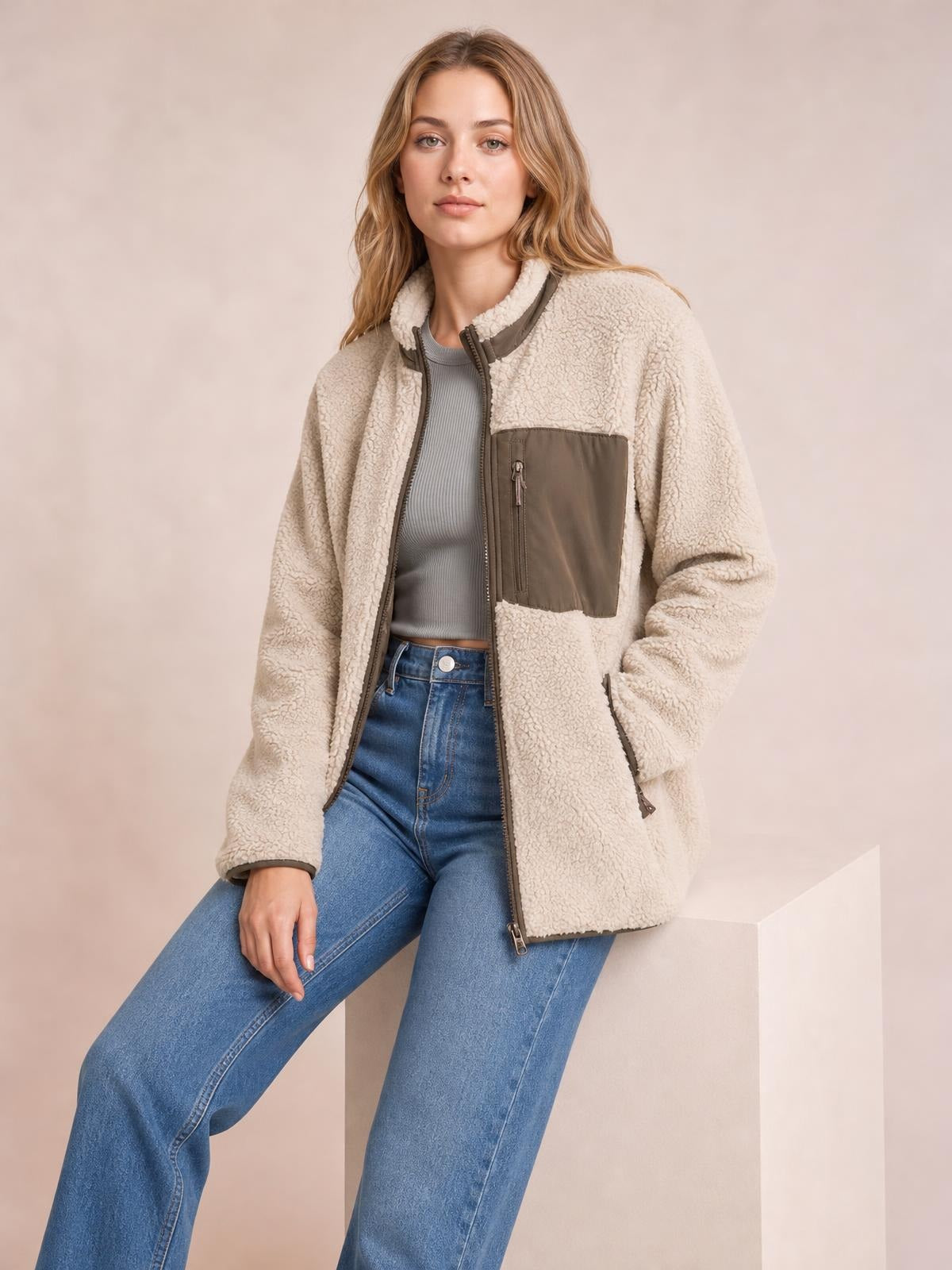 Model poses in a plush cream zip-up jacket with long sleeves, a high collar, brown contrast trim, and functional chest and side pockets.