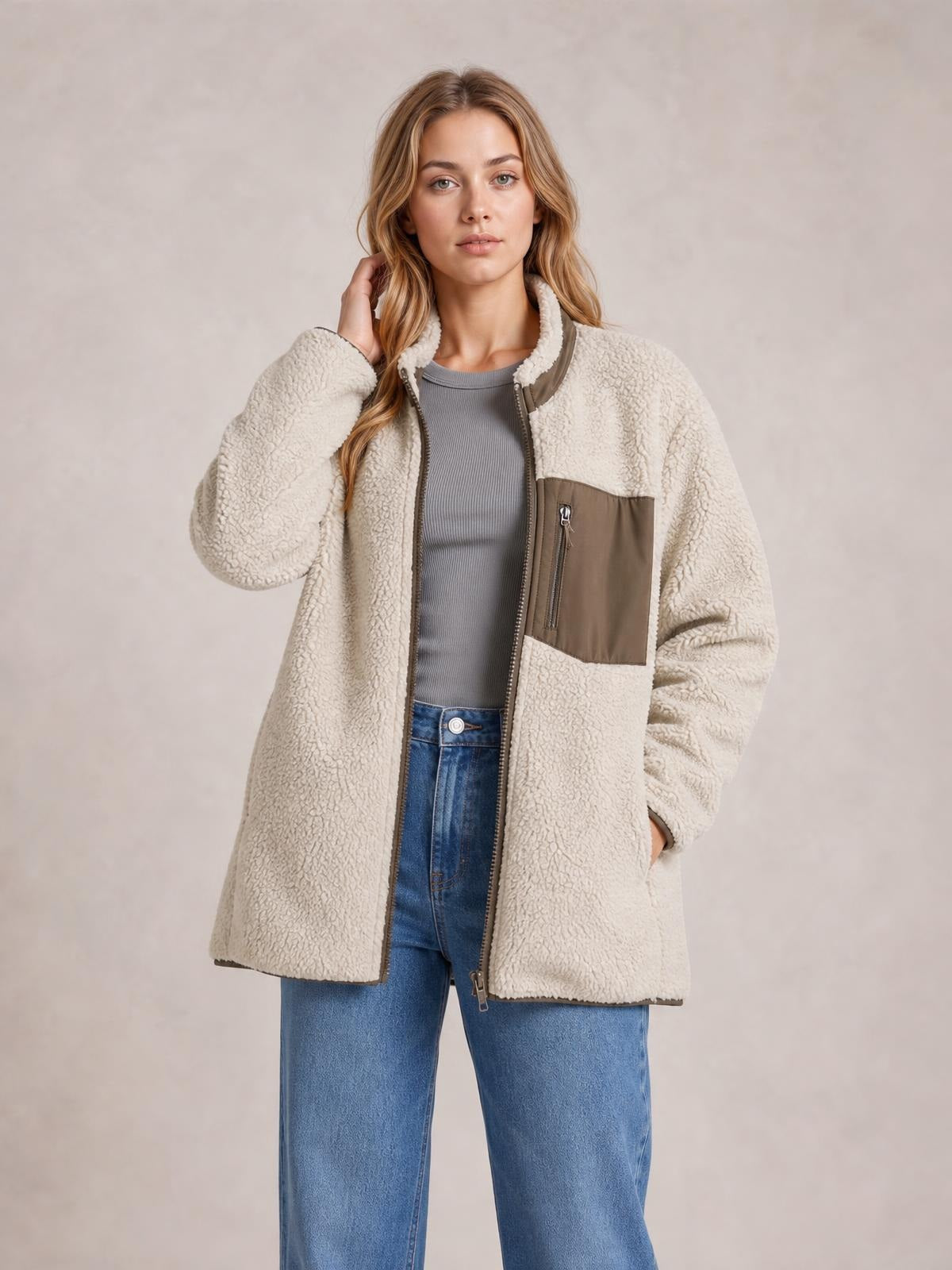 A woman models a cream plush zip-up jacket with long sleeves, a relaxed longline silhouette, contrast brown chest zip pocket, and one hand resting in a pocket.