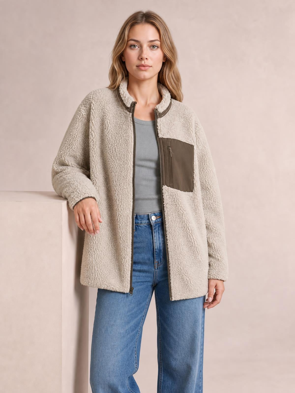 A model stands in a relaxed pose wearing an open cream plush zip-up jacket with long sleeves, a stand collar, brown chest pocket, and straight silhouette.